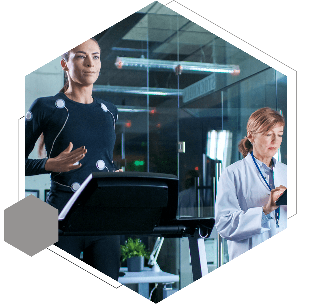 Person running on a treadmill wearing sensors, observed by a researcher in a lab coat taking notes.