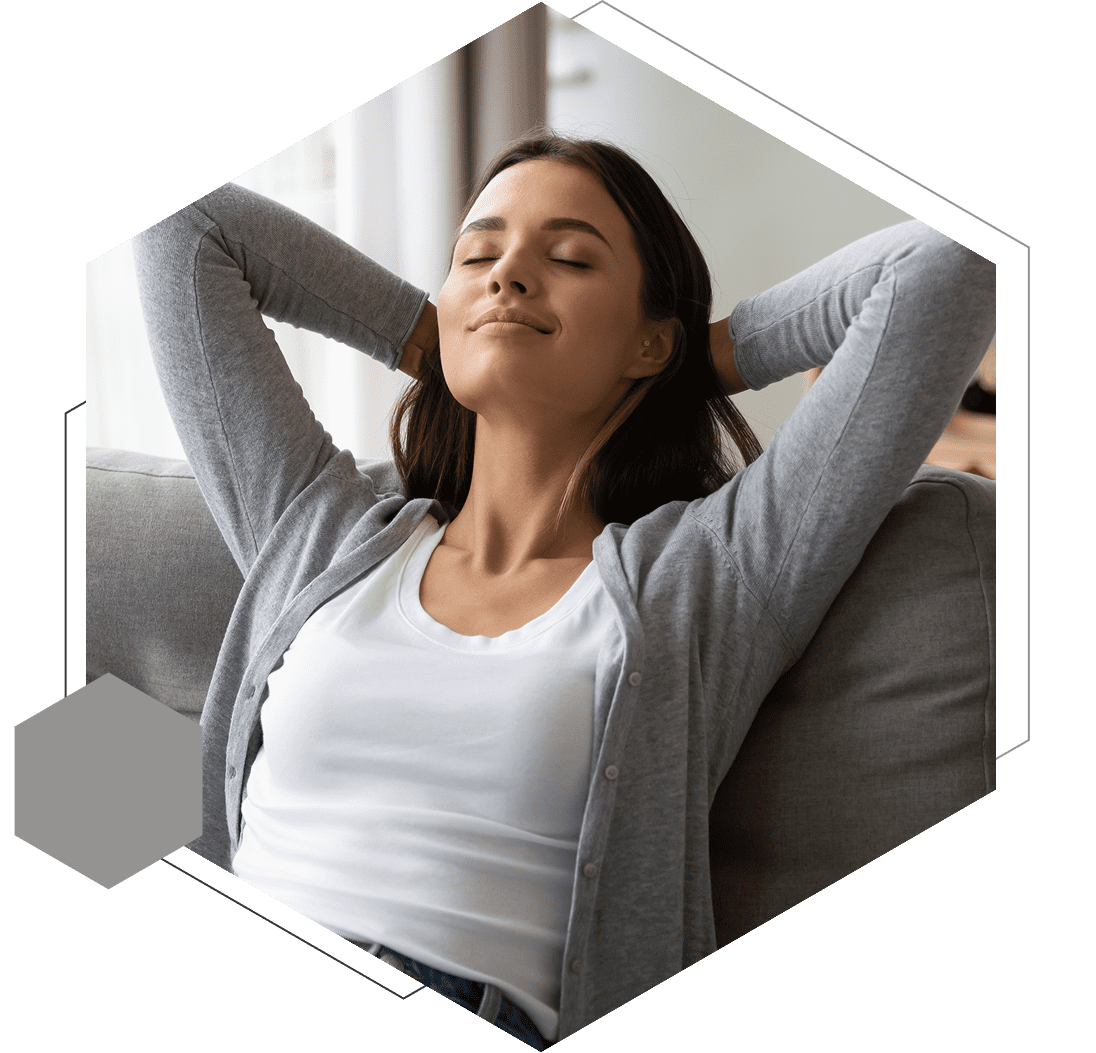 A woman sits on a couch with her hands behind her head, eyes closed, appearing relaxed. She is wearing a gray cardigan over a white top. The image is framed in a hexagon shape.