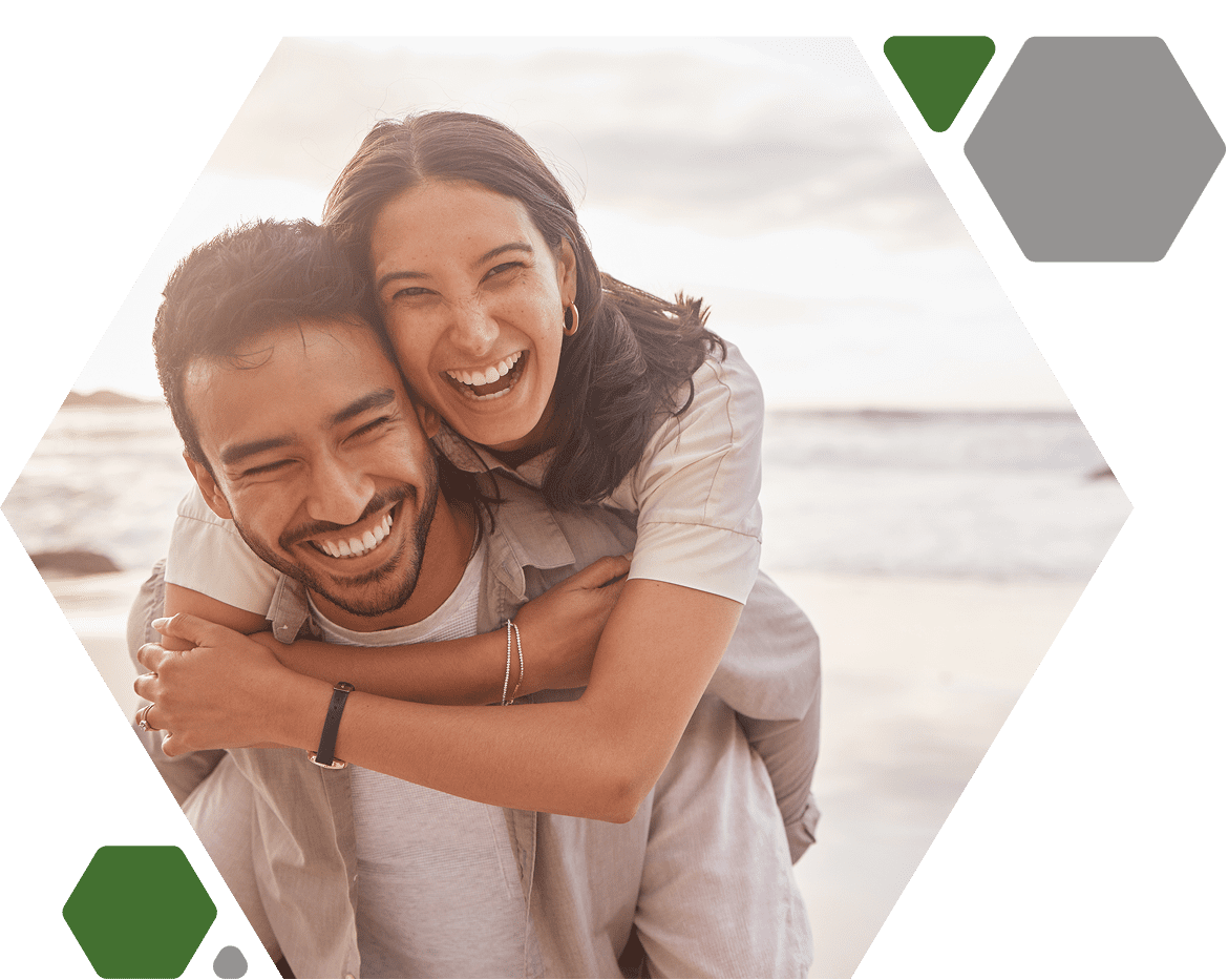 A couple smiling, with one person giving a piggyback ride to the other on a beach. Hexagonal frame overlays the image.
