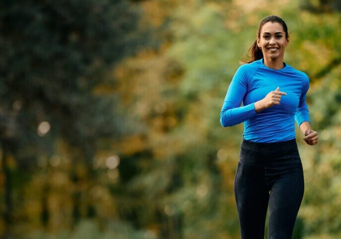 Woman jogging outdoors, wearing a blue top and black pants, with trees and greenery in the background.
